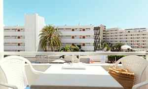 Näkymä parvekkeelta, jossa istuinryhmä, rakennuksia ja palmuja näkyvissä taustalla kirkkaan sinisen taivaan alla. (Tekoälyn luoma teksti)