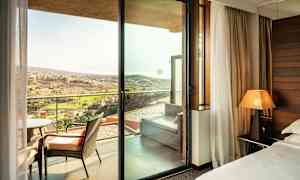 Hotellihuone, josta on näkymä lasioven kautta parvekkeelle, jossa on tuoleja ja kaukainen maisema ulkona. (Tekoälyn luoma teksti)