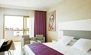 Hotellihuone, jossa on valkoiset vuodevaatteet ja violetti päiväpeite, kaksi tuolia pöydän ääressä ja parveke näkymällä. (Tekoälyn luoma teksti)