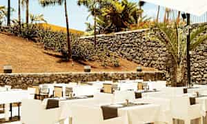 Ulkona sijaitseva ravintola, jossa on aseteltuja pöytiä ja tuoleja, ympäröity kivimuurilla ja palmuilla. (Tekoälyn luoma teksti)