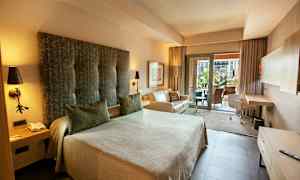 Hotellihuone, jossa on iso vuode, sohva ja kirjoituspöytä. Lisäksi on parveke näkymällä. (Tekoälyn luoma teksti)