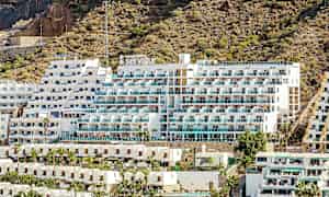 Kuva hotellista, jossa on valkoinen ulkoseinä ja parvekkeita, jotka sijaitsevat kivisen rinteen äärellä. (Tekoälyn luoma teksti)