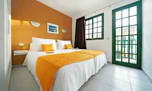 Makuuhuone, jossa on parivuode, valkoiset ja oranssit tekstiilit, sekä suuret vihreäreunaiset ikkunat. (Tekoälyn luoma teksti)