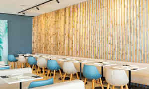 Ravintola, jossa vaaleat pöydät ja modernit tuolit puupaneelin edessä. (Tekoälyn luoma teksti)