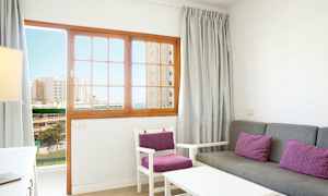 Olohuone, jossa harmaa sohva ja tuolit violetilla tyynyillä, ja puuovi lasi-ikkunoin, joka näyttää näkymän rakennuksiin ja merelle. (Tekoälyn luoma teksti)