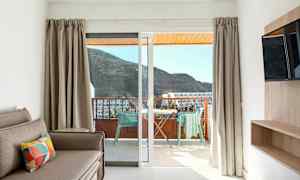 Näkymä hotellihuoneen sisätiloista parvekkeelle, jossa on pöytä ja tuolit, sekä taustalla vuoristomaisema. (Tekoälyn luoma teksti)