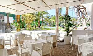 Ulkoterassi, jossa valkoisia pöytiä ja tuoleja, näkymä vehreään puutarhaan. (Tekoälyn luoma teksti)