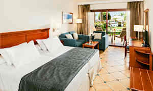 Hotellihuone, jossa leveä sänky, oleskelualue ja terassi, josta on näkymä vehreään puutarhaan. (Tekoälyn luoma teksti)