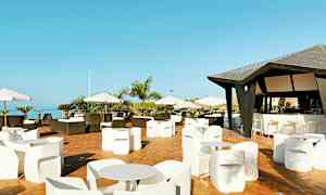 Terassi, jossa on pöytiä ja tuoleja sekä katoksella varustettu baari. Näkymää koristavat aurinkovarjot ja taivasta sekä puita. (Tekoälyn luoma teksti)