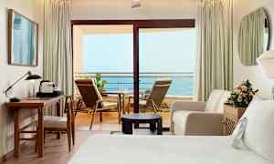 Hotellihuone, jossa on parveke merinäköalalla. Huoneessa on kirjoituspöytä, tuoli, sohva, sänky ja kukkia. (Tekoälyn luoma teksti)