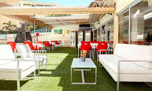 Ulkoterassi, jossa valkoisia sohvia, punaisia tuoleja ja pöytiä, aseteltu tekonurmelle ja pergolan alle. (Tekoälyn luoma teksti)