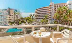 Näkymä aurinkoiselta parvekkeelta, jossa on pöytä ja tuolit, hotellirakennus ja uima-allas näkyvät taustalla. (Tekoälyn luoma teksti)