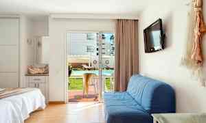 Hotellihuone, jossa on pedattu sänky, sininen sohva, seinään kiinnitetty televisio ja lasiovi, joka johtaa terassille, jossa on pöytä ja tuolit. Näkymä näyttää ulkouima-altaan ja aurinkotuoleja. (Tekoälyn luoma teksti)