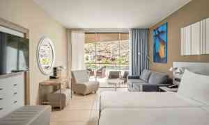 Hotellihuone, jossa kaksi yhden hengen sänkyä, sohva ja istuimet. Lasiovet johtavat terassille näköalalla. (Tekoälyn luoma teksti)