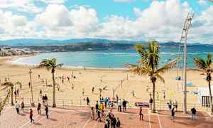 Ranta, jossa ihmiset pelaavat lentopalloa ja kävelevät rantakadulla. Meri ja vuoret näkyvät taustalla. (Tekoälyn luoma teksti)