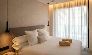 Hotellihuone, jossa on suuri sänky, valkoiset lakanat ja tyynyt. Sängyllä on rullattuja pyyhkeitä. Seinät ja verhot ovat vaaleita ja huoneessa on riippuvalaisimet. (Tekoälyn luoma teksti)