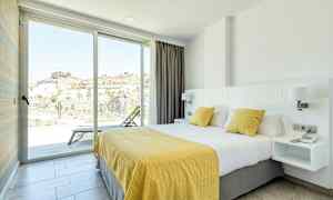 Hotellihuone, jossa on suuri sänky, joka on peitetty valkoisilla ja keltaisilla vuodevaatteilla. Huone on valoisa ja avara, ja siitä on lasiovet parvekkeelle, josta on näkymä vuoristoiseen alueeseen. (Tekoälyn luoma teksti)