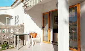 Terassi, jossa on valkoinen rakennus, pöytä ja tuoli sekä taloon johtava ovi. (Tekoälyn luoma teksti)