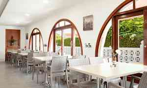 Ravintola, jossa kaarevat ikkunat ja pöytiä tuoleineen. Ulkona näkyy kasveja ikkunan läpi. (Tekoälyn luoma teksti)