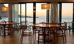 Ravintola, jossa puiset tuolit ja pöydät, näkymä merelle ja auringonlaskuun. (Tekoälyn luoma teksti)