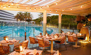 Ulkoterassi, jossa katetut pöydät ja valoköynnöksiä, sijaitsee altaan lähellä ja palmuja ympäröivät sen. (Tekoälyn luoma teksti)