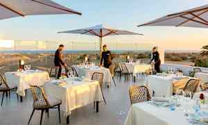 Ulkoterassi, jossa katetut pöydät ja aurinkovarjoja, taustalla näkymä hiekkadyyneille. (Tekoälyn luoma teksti)