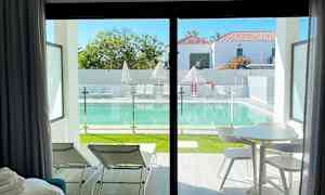 Näkymä huoneesta, jossa on tuoleja ja pöytä, uima-altaalle, jonka äärellä aurinkotuoleja ja varjoja. (Tekoälyn luoma teksti)