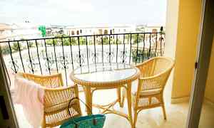 Kaksi rottinkituolia ja pyöreä lasipöytä parvekkeella, näkymä kivirakennuksiin ja tenniskenttään. (Tekoälyn luoma teksti)