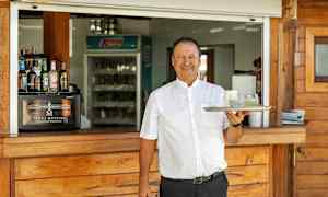 Henkilö pitää tarjotinta, jossa on kaksi lasia, puisen tarjoilutiskin edessä. (Tekoälyn luoma teksti)