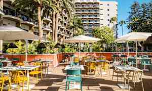 Kuvassa on ulkona sijaitseva ravintola, jossa on värikkäitä tuoleja, pöytiä ja varjoja, hotellin parvekkeiden edessä. (Tekoälyn luoma teksti)