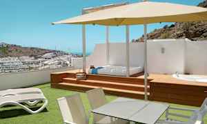 Terassi, jossa aurinkotuoleja, pöytä, varjo sekä näkymä mäkiseen ympäristöön rakennuksineen. (Tekoälyn luoma teksti)