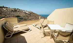 Terassi, jossa aurinkotuoleja sekä pöytä ja tuoleja, näkymä merelle. (Tekoälyn luoma teksti)