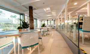 Ravintolan sisustus, jossa on valkoiset tuolit ja pöydät, lasiseinät ja näkymä ulkona oleviin viheralueisiin. (Tekoälyn luoma teksti)