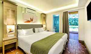 Moderni hotellihuone, jossa kaksi yhden hengen vuodetta, vihreät sängynpeitot ja valkoinen sisustus, näkymä parvekkeelle ja kylpyhuoneeseen. (Tekoälyn luoma teksti)