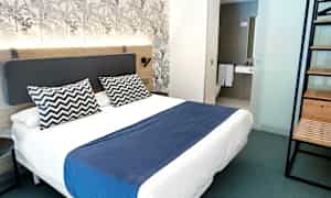 Moderni hotellihuone, jossa on parivuode, sininen päiväpeitto ja kaksi kuviollista tyynyä. (Tekoälyn luoma teksti)