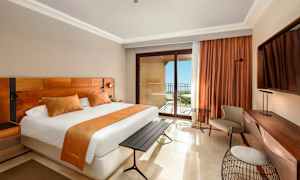 Hotellihuone, jossa on suuri sänky, oransseja sisustusyksityiskohtia, kirjoituspöytä ja parveke merinäkymällä. (Tekoälyn luoma teksti)