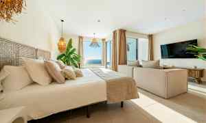 Kuva valoisasta ja modernista makuuhuoneesta, jossa on suuri sänky, sohva, televisio ja laaja merinäköala suurien ikkunoiden läpi. (Tekoälyn luoma teksti)