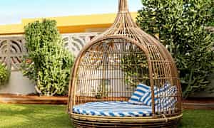 Rottinkikaluste, jossa on katos ja sinivalkoiset raidalliset tyynyt, sijaitsee nurmikolla vihreiden pensaiden vieressä. (Tekoälyn luoma teksti)