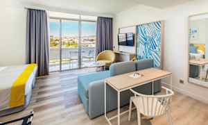 Hotellihuone, jossa on sänky, kirjoituspöytä, sohva, televisio ja lasiovet, jotka johtavat parvekkeelle. (Tekoälyn luoma teksti)