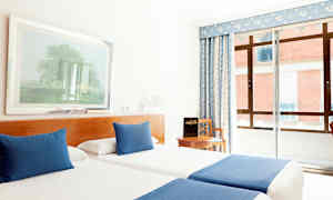 Hotellihuone, jossa on kaksi sinisillä peitoilla ja tyynyillä varustettua yhden hengen sänkyä, tuoli pöydän ääressä ja ikkuna sinisellä verholla. (Tekoälyn luoma teksti)