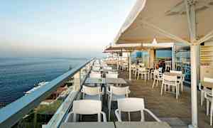 Kuva ulkoterassista, jossa on pöytiä, tuoleja ja varjoja, merinäköalalla. (Tekoälyn luoma teksti)