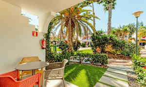 Terassi, jossa pöytä ja värikkäitä tuoleja, rajattuna kaarimuodolla, joka avautuu puutarhaan palmuineen ja rakennuksineen taustalla. (Tekoälyn luoma teksti)