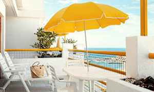 Valkoinen terassi, jossa on tuolit, pöytä, keltainen aurinkovarjo ja merinäkymä. (Tekoälyn luoma teksti)