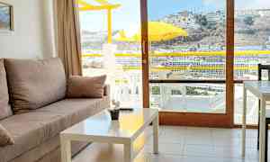 Huone, jossa on sohva ja sohvapöytä, avautuu parvekkeelle, jossa on keltainen aurinkovarjo ja näkymä kumpuilevaan alueeseen. (Tekoälyn luoma teksti)