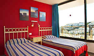 Kaksi yhden hengen sänkyä raidallisilla vuodevaatteilla ja punainen seinä, jossa on kuvia, sekä suuri ikkuna näkymällä rakennuksiin. (Tekoälyn luoma teksti)