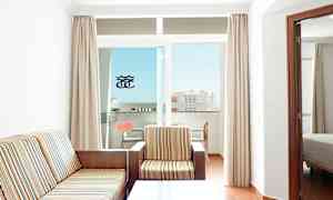 Huone, jossa on raidallisella kankaalla verhoiltu sohva ja nojatuoli, sohvapöytä ja parveke, josta näkyy rakennus. (Tekoälyn luoma teksti)