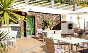 Terassi, jossa on pöytiä, tuoleja ja aurinkovarjo, lähellä rakennusta, jossa on kivinen julkisivu. (Tekoälyn luoma teksti)