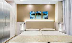 Kuvassa on moderni hotellihuone, jossa on parivuode, tyylikkäät yöpöydät ja seinäkoriste, johon kuuluu kolme trooppista rannalla tehtyä kuvaa. (Tekoälyn luoma teksti)