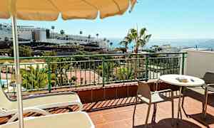 Terassi, jossa on pöytä, tuolit ja aurinkotuolit. Taustalla näkyy palmuja, rakennuksia ja meri. (Tekoälyn luoma teksti)