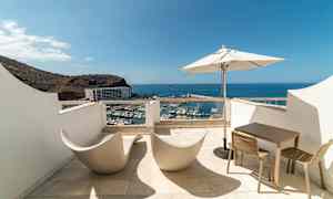 Terassi, jolta on näkymä satamaan ja merelle, varustettuna moderneilla tuoleilla, pöydällä ja aurinkovarjolla. (Tekoälyn luoma teksti)